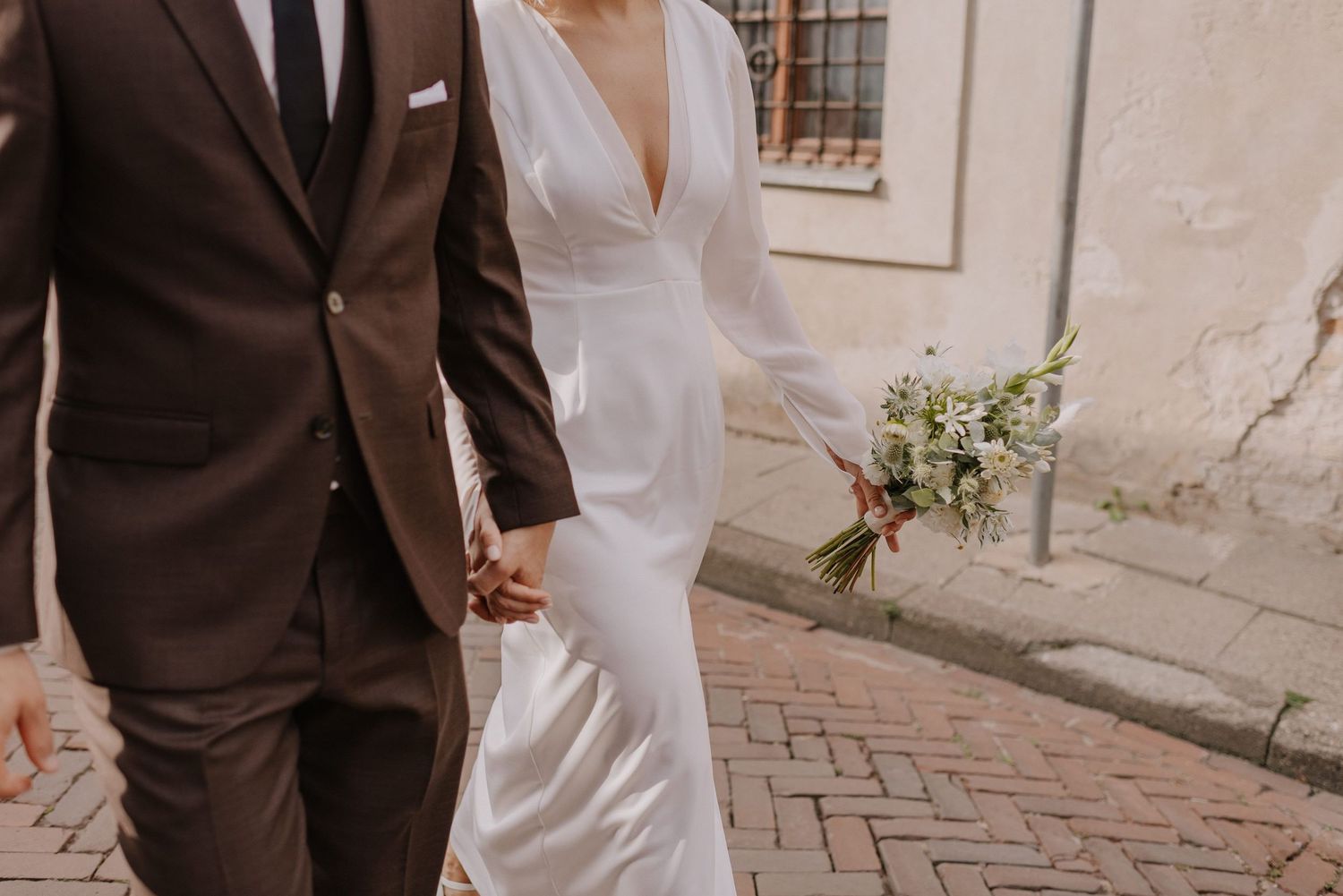 Elegant wedding couple walking together while holding bouquet along stone walls.