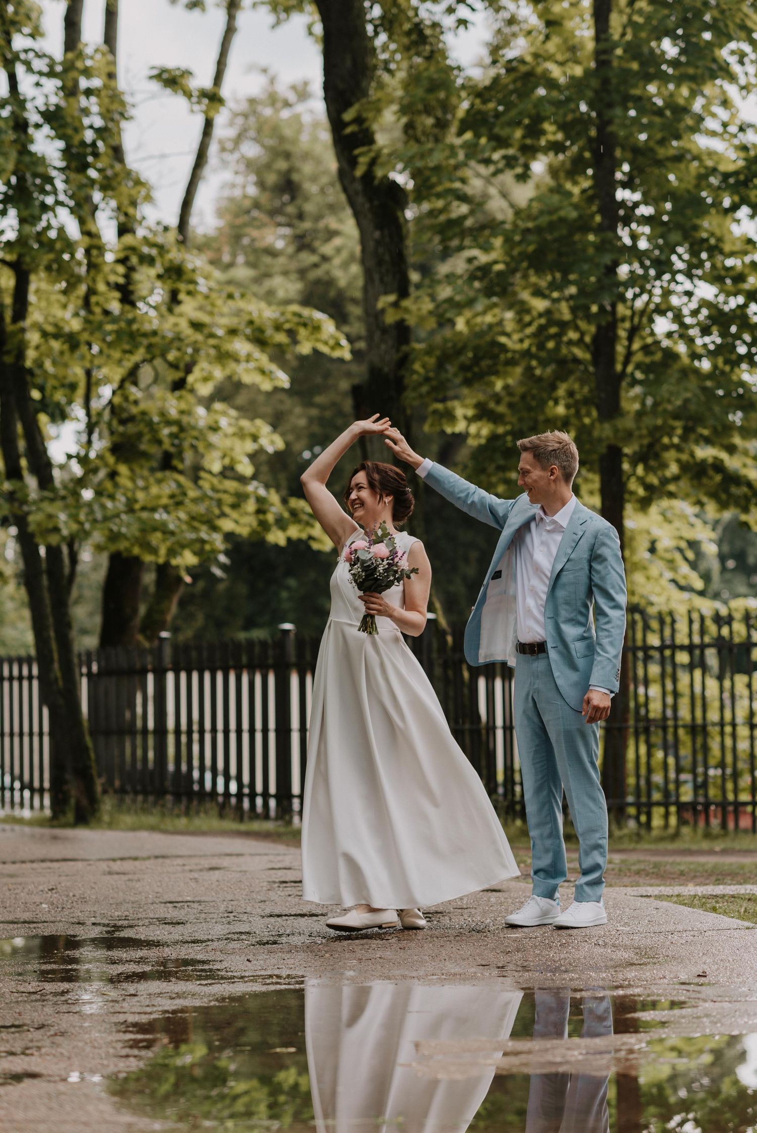 Dancing couple in wedding attire twirl playfully in a park after rainfall.