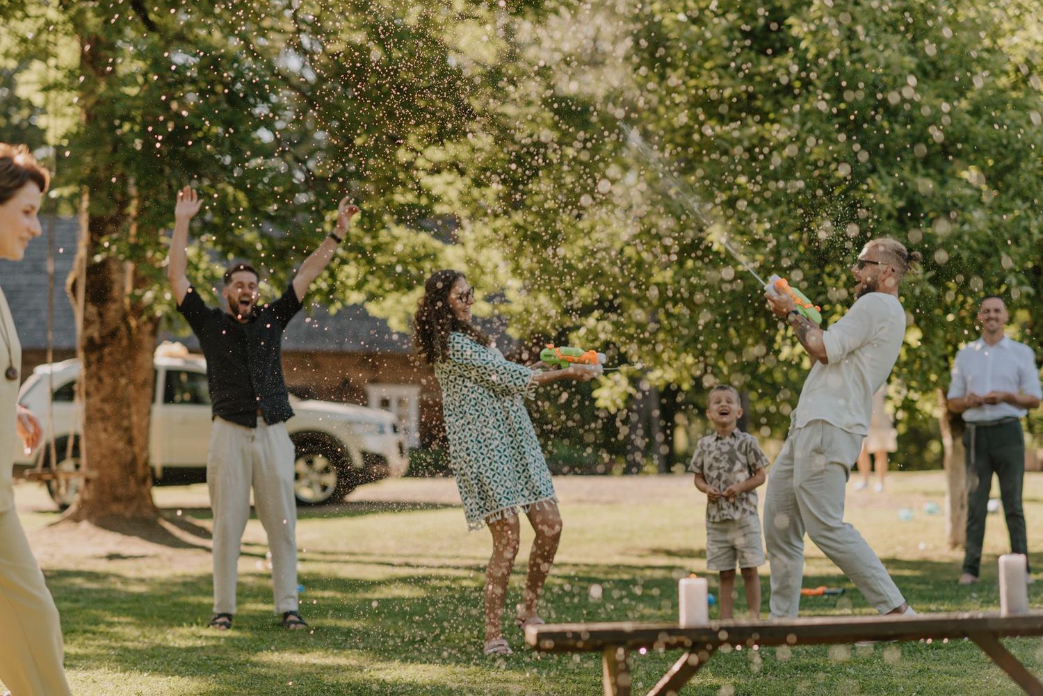 Wedding celebration scene with guests mingling under large trees in a garden setting.