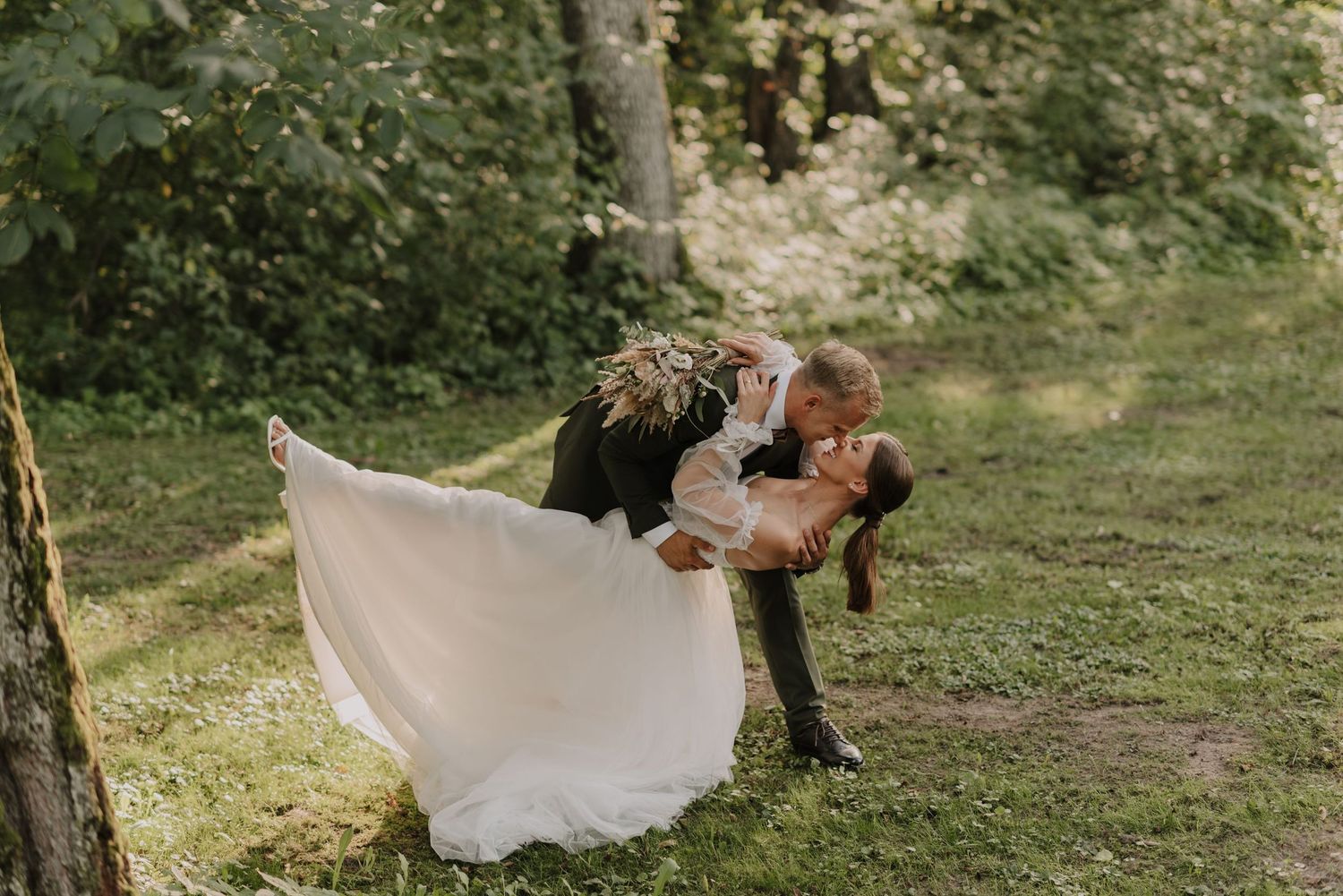 A romantic dip pose between newlyweds in a sun-dappled forest setting.