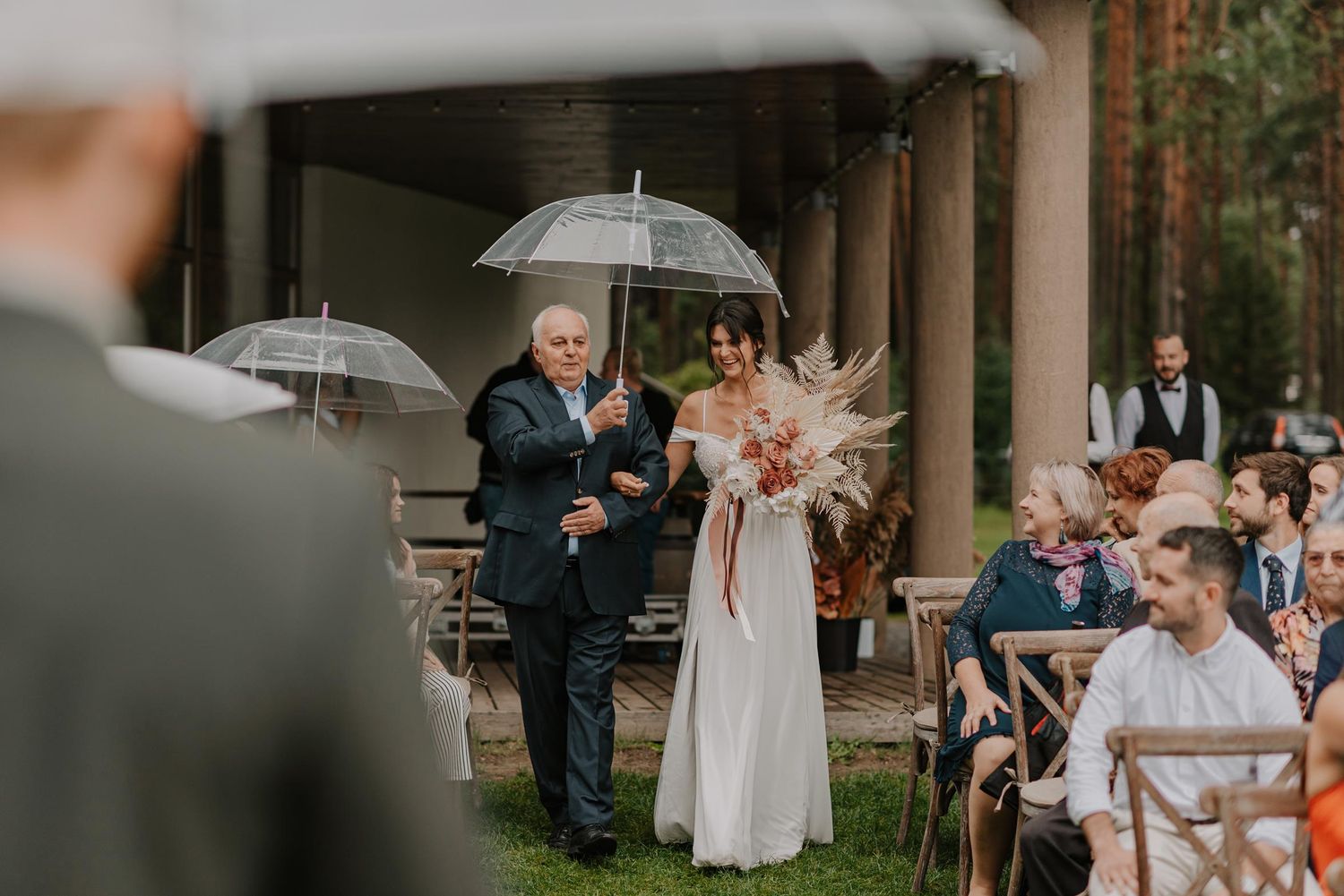 Guests with umbrellas gather under a covered outdoor venue during a rainy wedding celebration.
