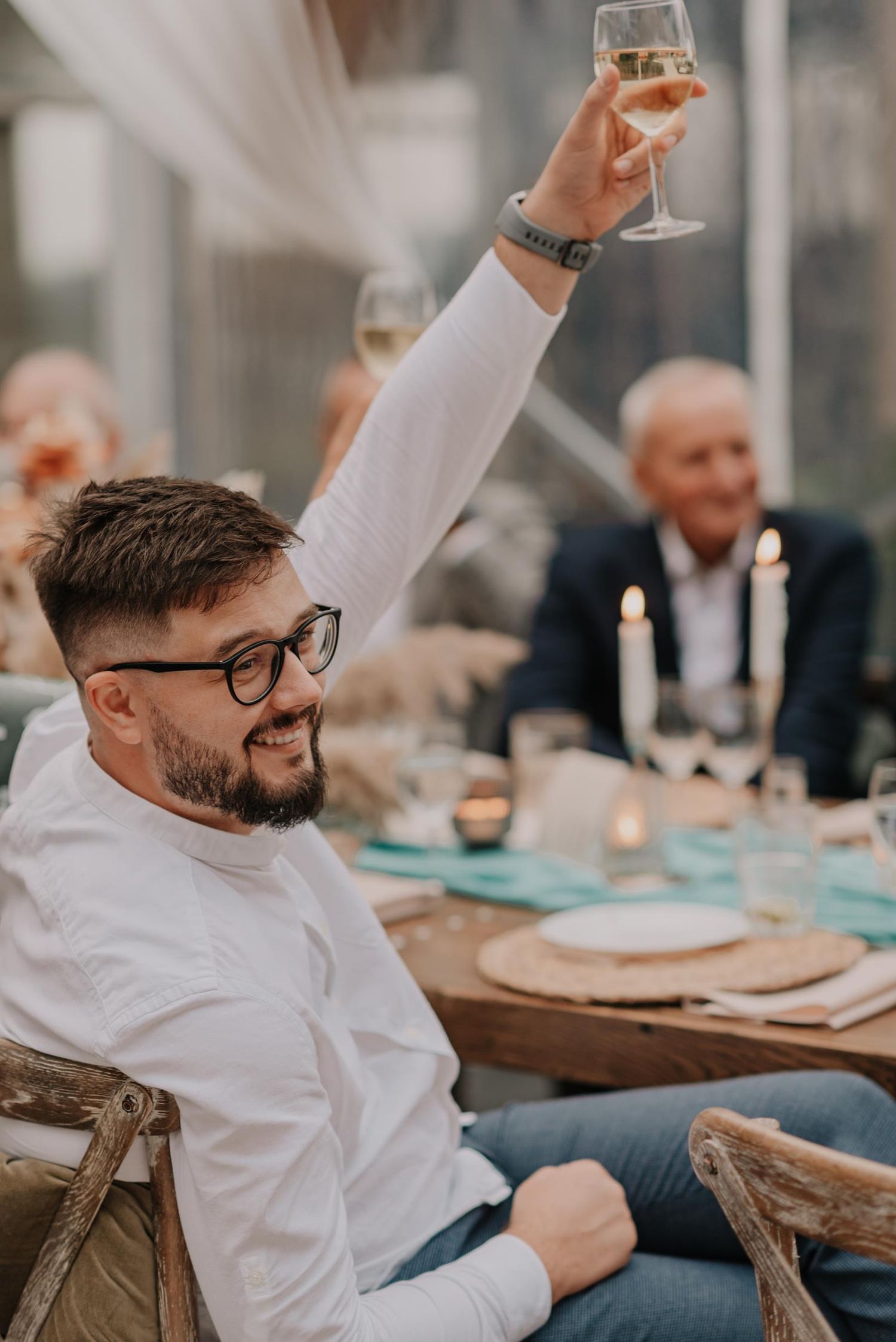 Guest in white shirt and glasses raises wine glass in celebratory toast at reception.
