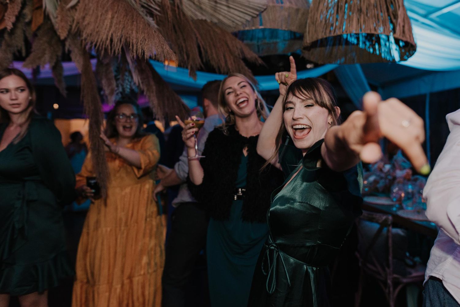 Wedding guests dance joyfully at a reception under a thatched roof with colorful lighting and festive atmosphere.