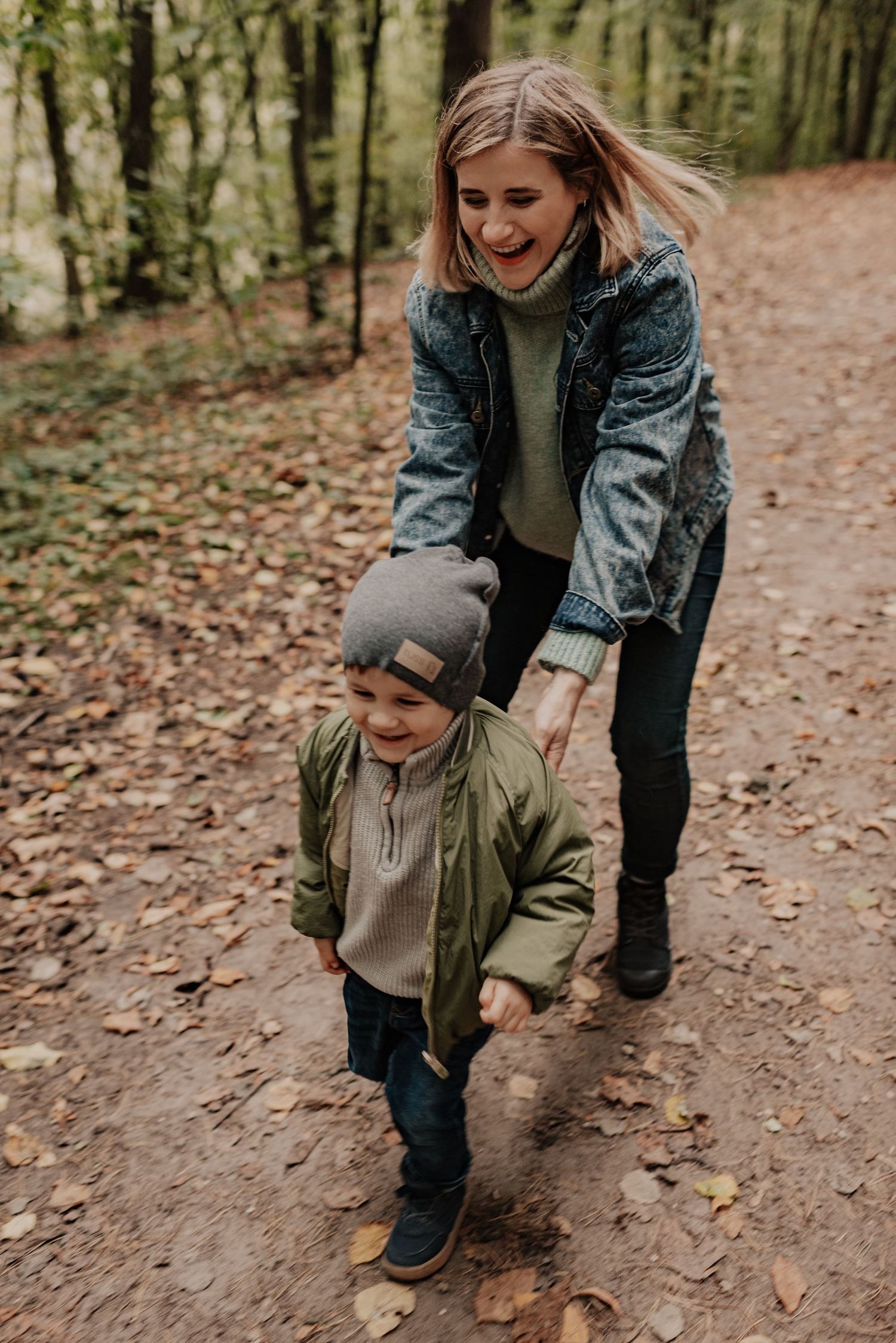 Parent and child exploring autumn forest trail together while looking at fallen leaves on ground.
