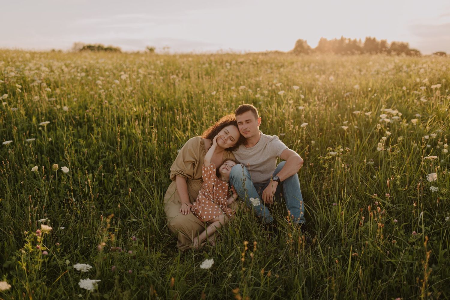 Family cuddles together while sitting in tall grass during sunset.