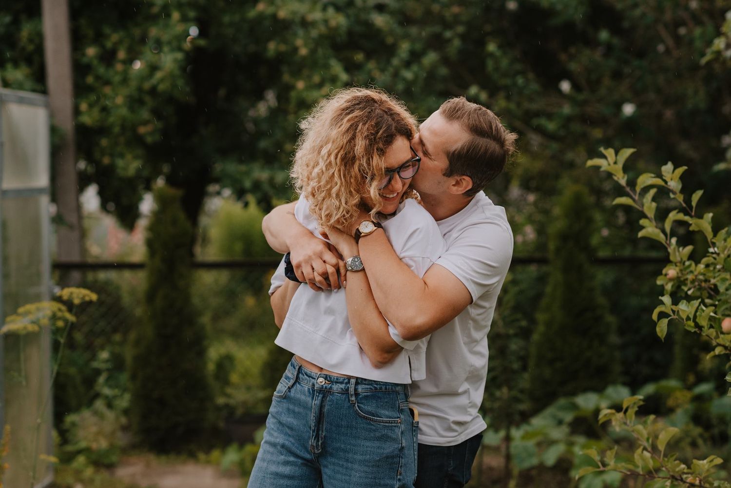 A couple embraces each other outdoors against a backdrop of lush green foliage.