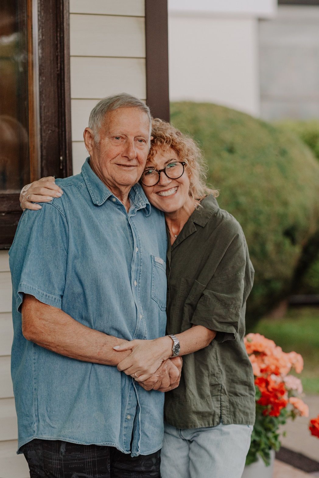 A couple embraces and smiles together on their front porch with red flowers in the background.
