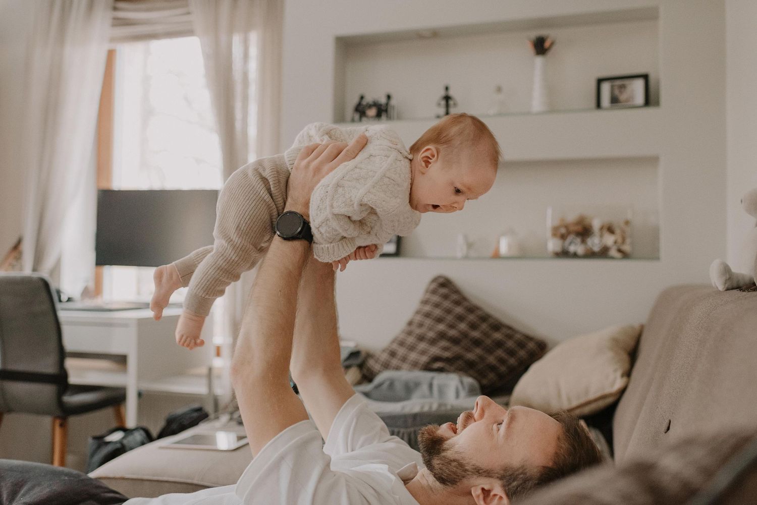 Parent lifting infant high in cozy living room with built-in shelves in background.