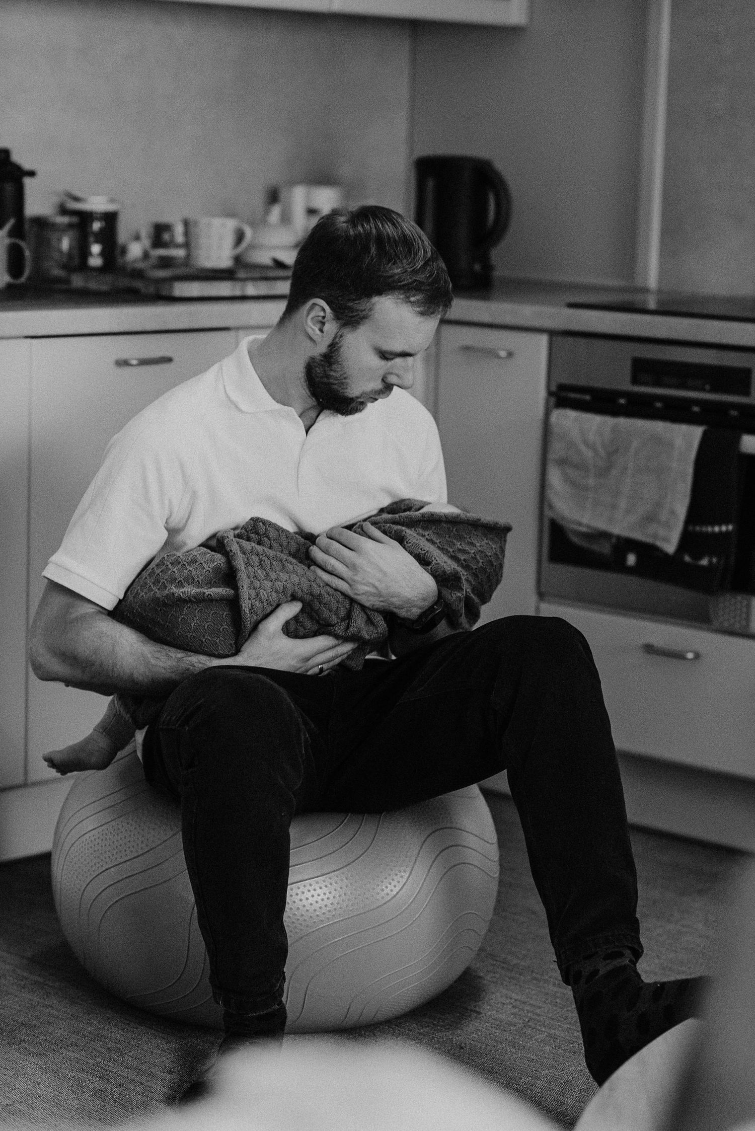 Parent sitting on exercise ball holding swaddled infant in modern kitchen.