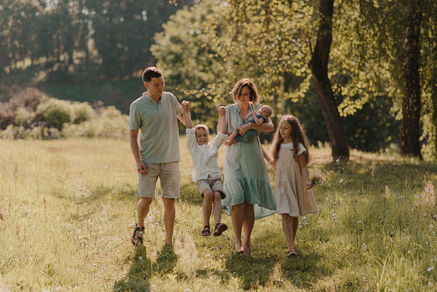 Family walking together through a sun-lit meadow surrounded by trees.