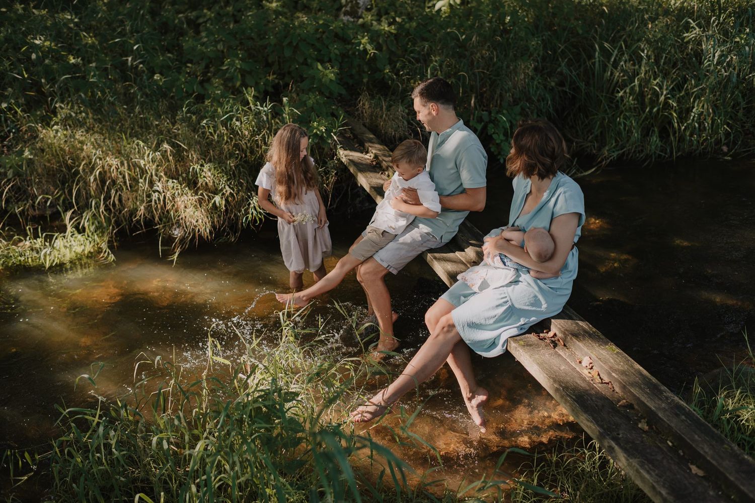 Peaceful outdoor scene with people sitting by a creek in dappled sunlight surrounded by greenery.
