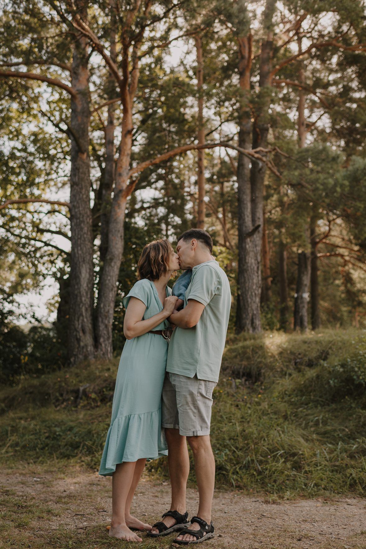 A romantic couple shares tender moments in a wooded setting wearing mint green and beige outfits at sunset.