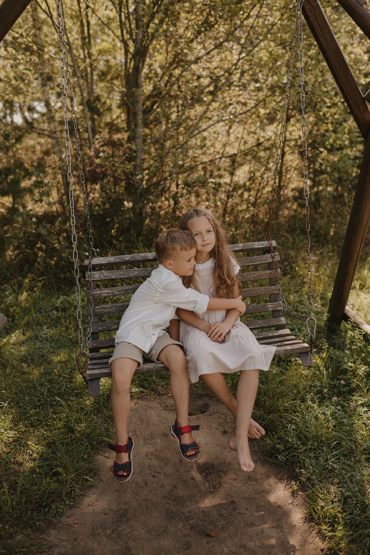 Siblings share sweet moments while sitting on a wooden swing in a forest setting.