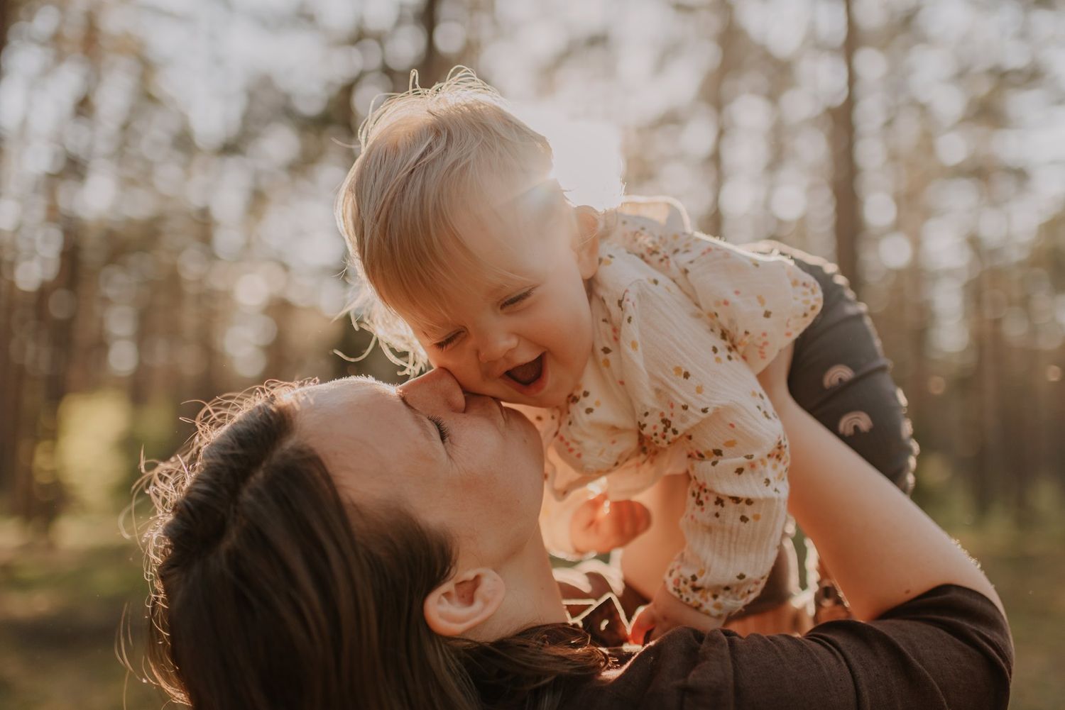 Parent and child share loving embrace in sunlit forest with lens flare creating magical atmosphere.