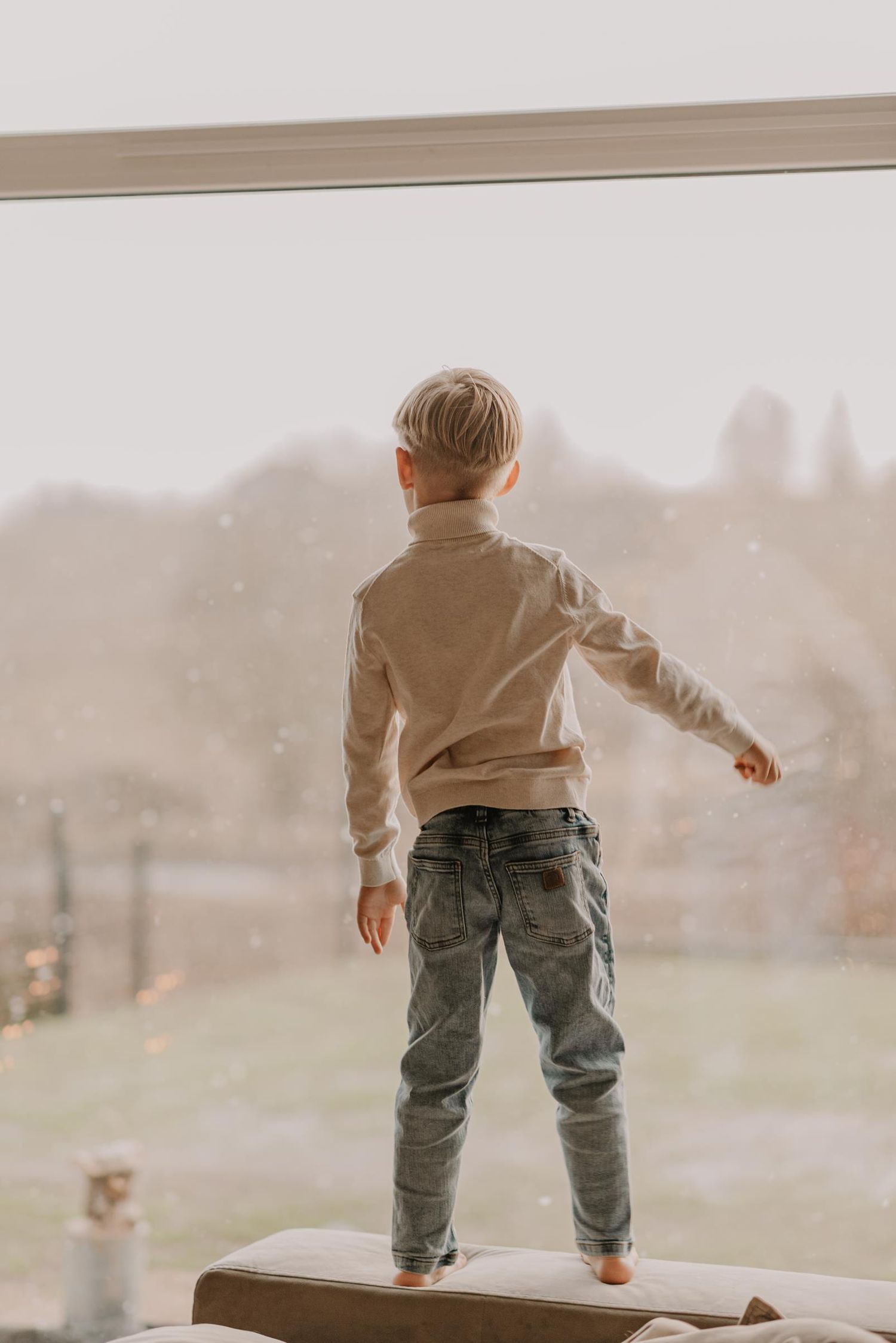 Child in tan jacket and camo pants jumping and playing on windowsill at home in moody lighting.