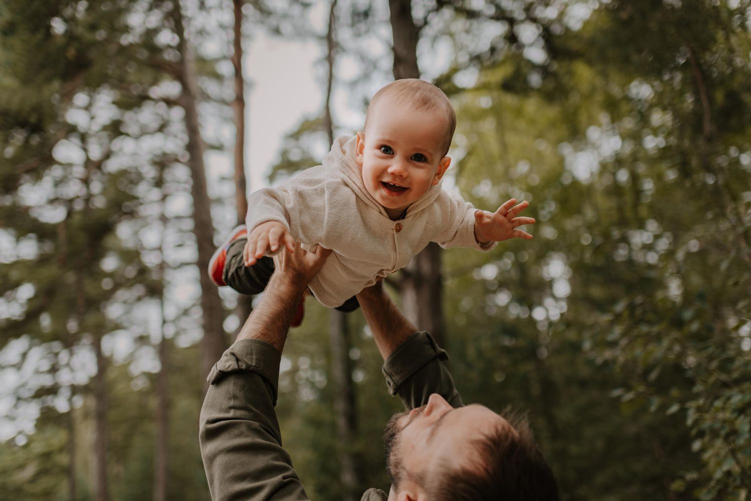 Baby being lifted playfully in air against forest backdrop.