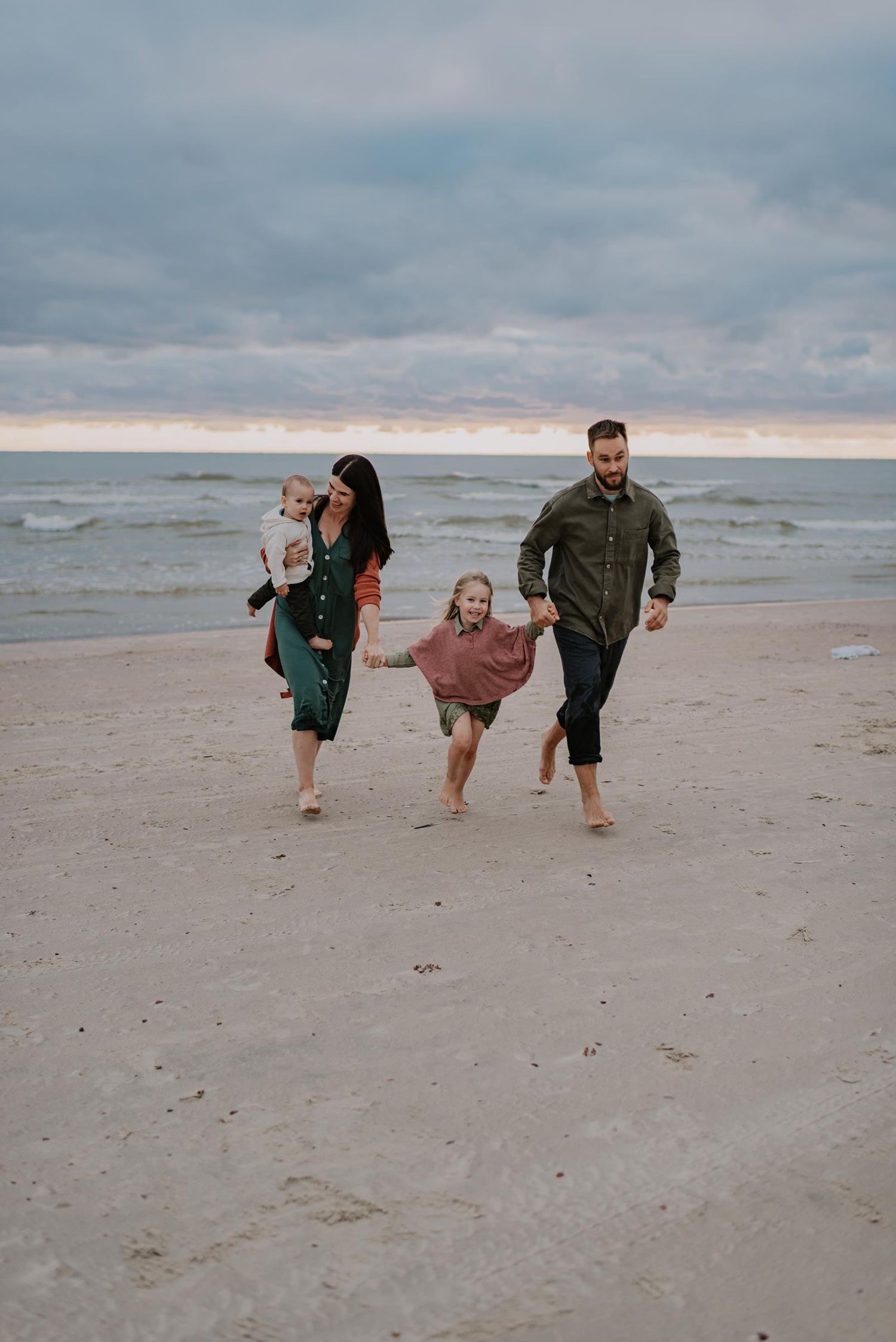 Family runs and plays together on sandy beach during overcast day.