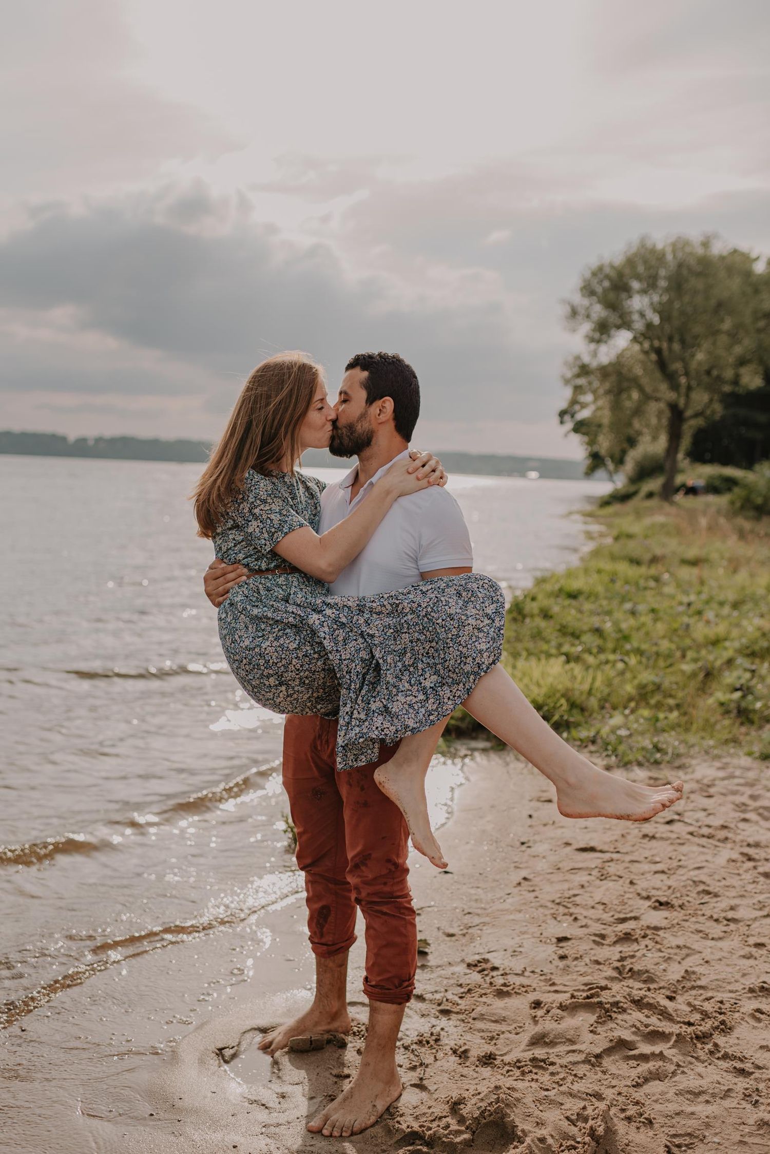 Romantic couple embracing on a beach shore with water and cloudy sky in background.