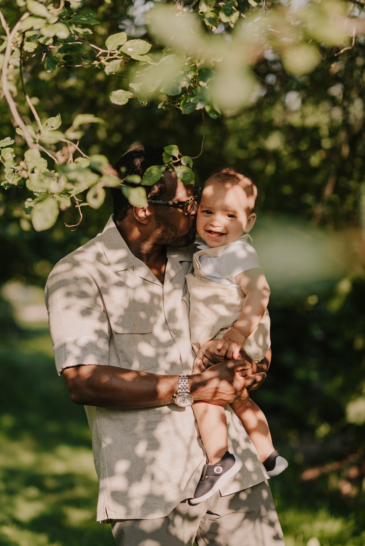Sunlight dapples through trees as a tender family moment unfolds in a garden setting.