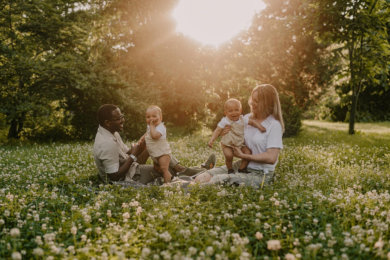 Family enjoying golden hour in a blooming meadow field with white flowers and sunlight streaming through trees.
