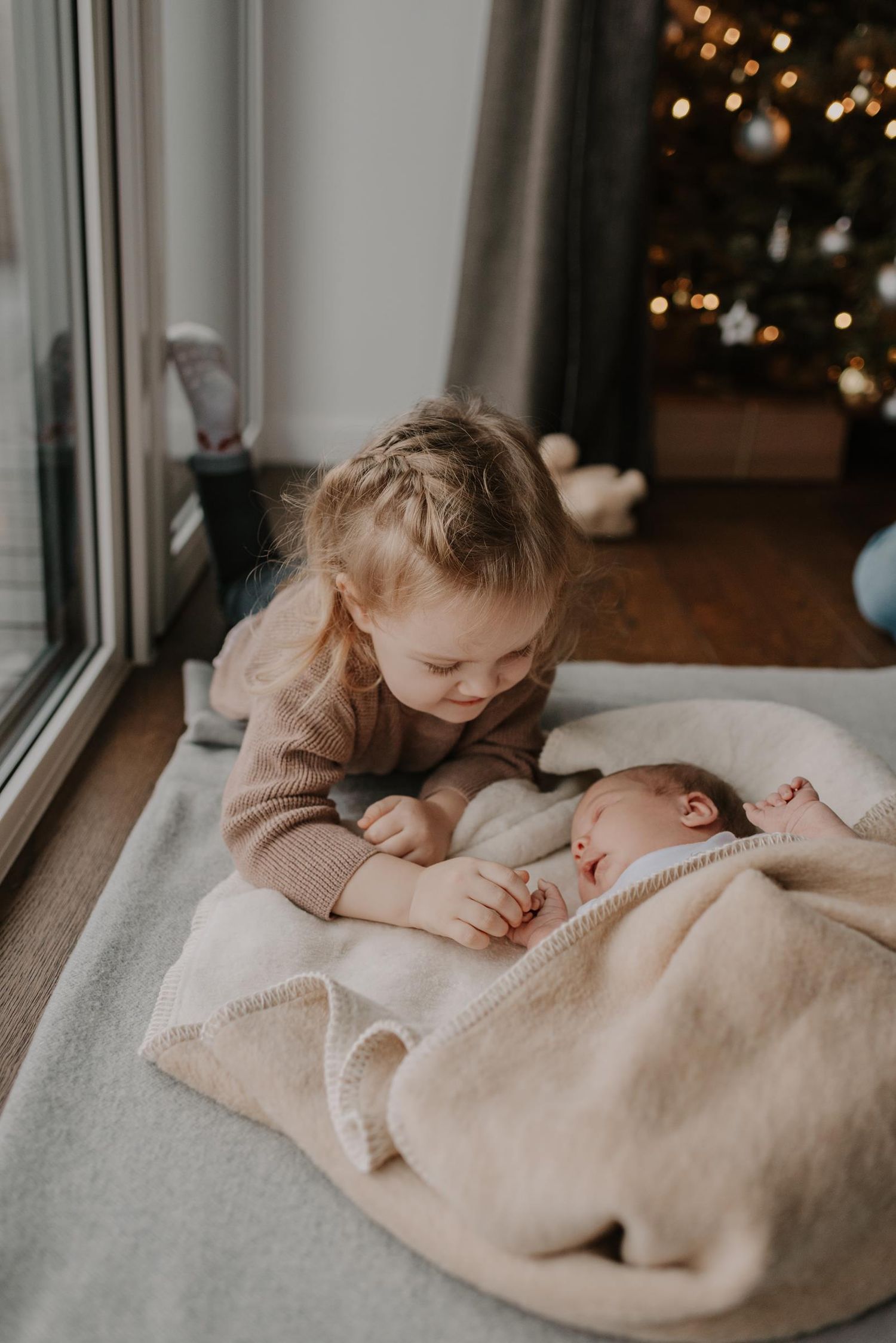 Siblings bonding with newborn baby by window with Christmas lights in background.