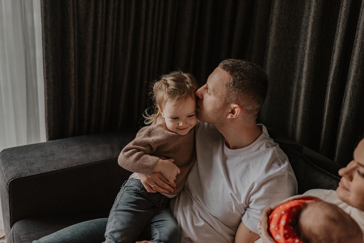Family sharing intimate moment with baby on dark couch.