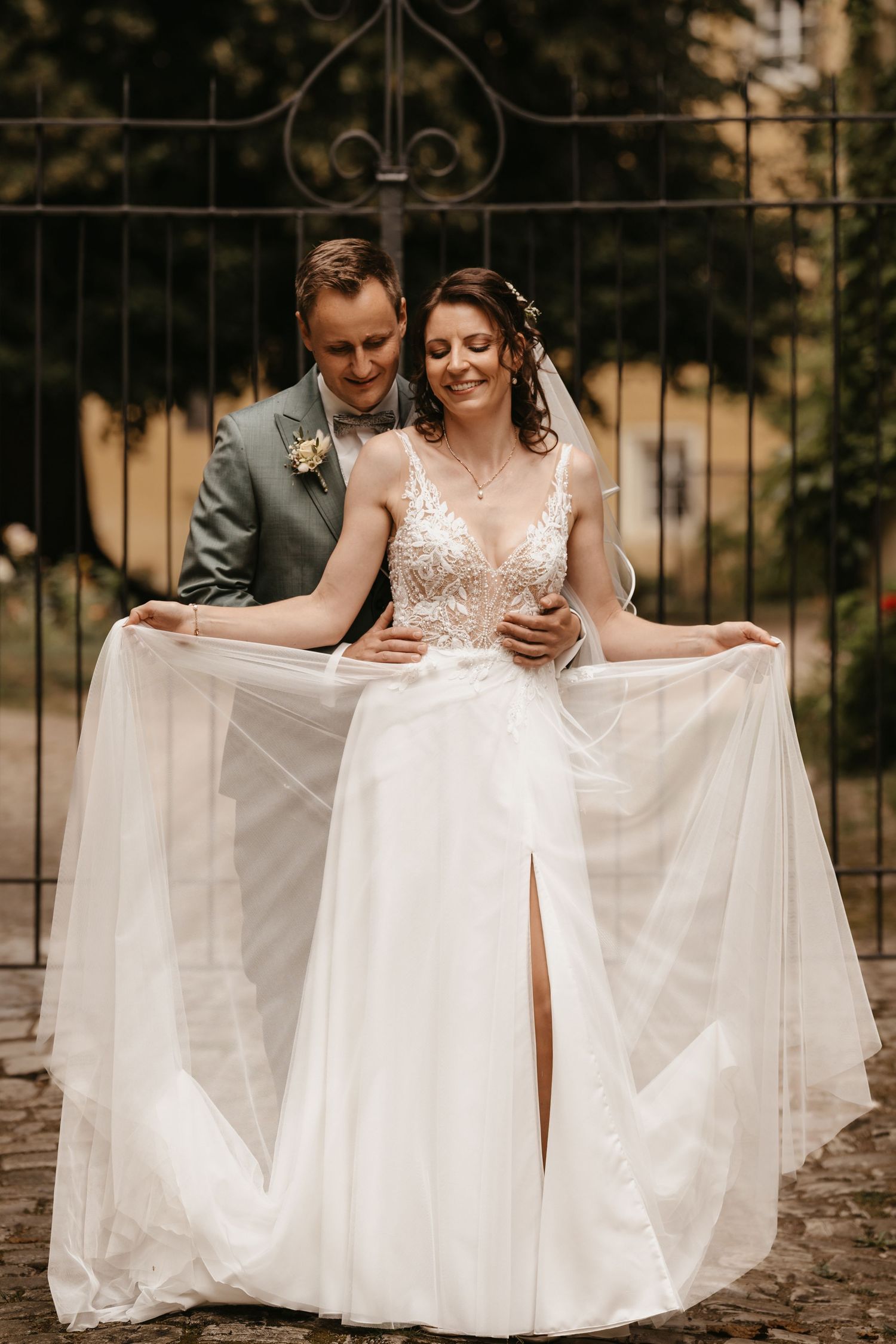 A couple in wedding attire sits together on stone steps while holding out the flowing white tulle skirt of the dress.