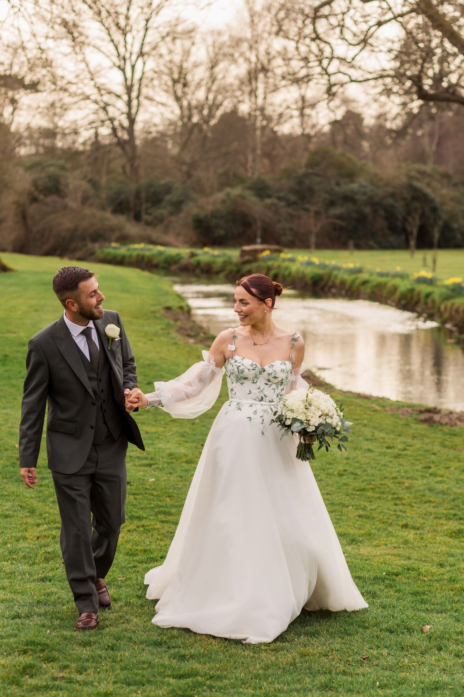A couple in wedding attire poses for photos in front of a brick building and garden landscape.