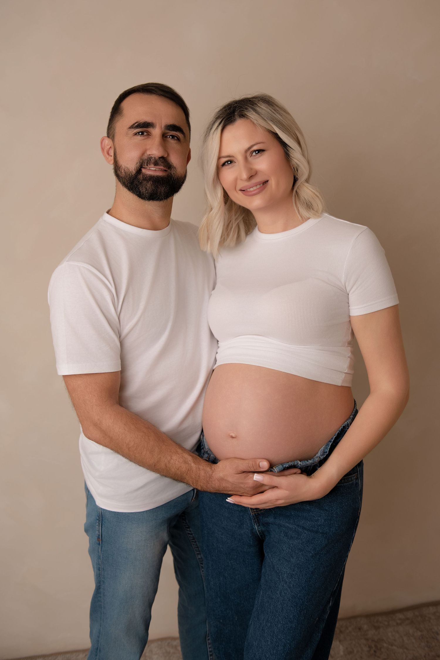 A couple in white t-shirts and jeans pose for a maternity photo series against a neutral backdrop.