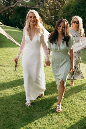 Three women walking on a sunny lawn, one wearing a white wedding dress and another in a sage green dress.