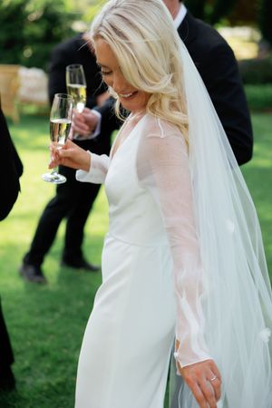 Bride in white dress with flowing veil walking on green lawn during wedding celebration.