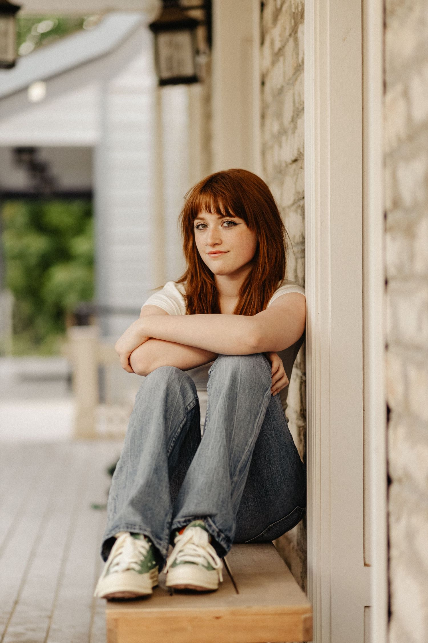 Person in jeans sits on wooden ledge against white columns in contemplative pose wearing casual sneakers.