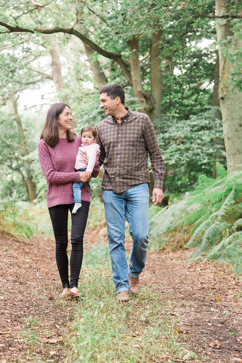 A couple walks together with their baby along a wooded trail covered in fallen leaves on an autumn day.