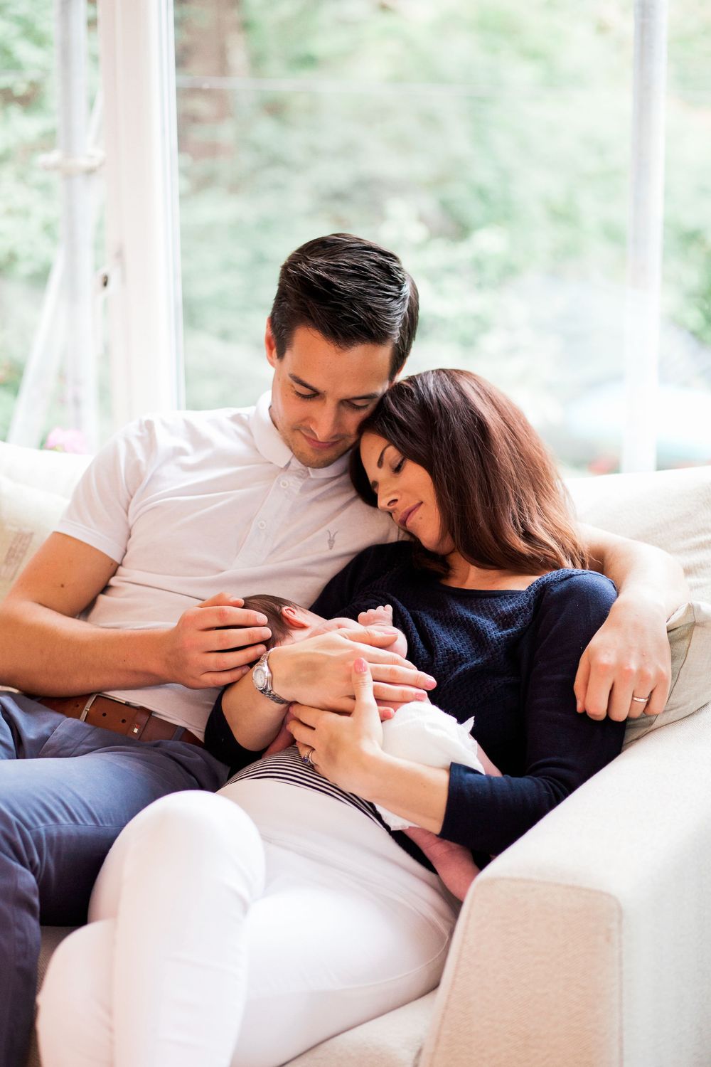 A couple relaxes together on a white couch while looking at a their newborn baby.