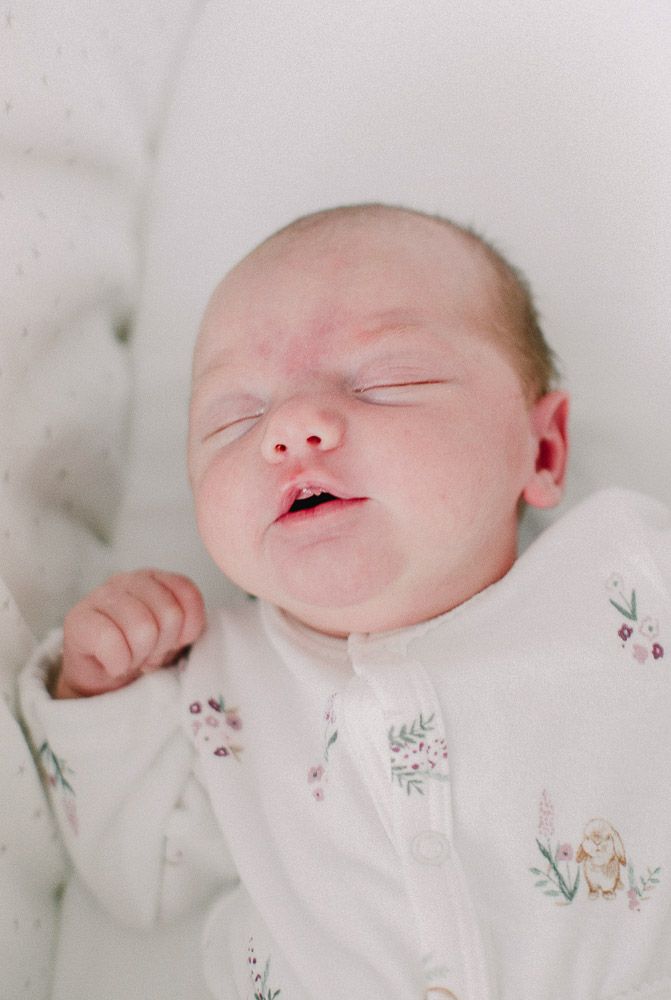 A sleeping newborn baby wearing a white onesie with delicate floral embroidery lies on soft white bedding.