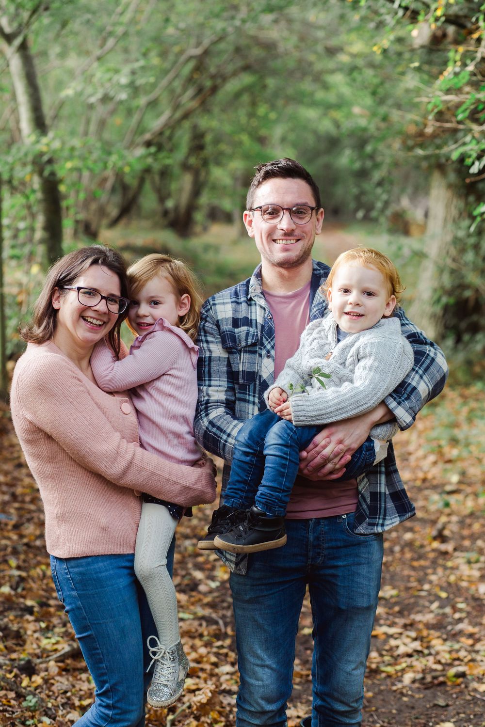 A family of four poses together on a leaf-covered path in an autumn forest setting.
