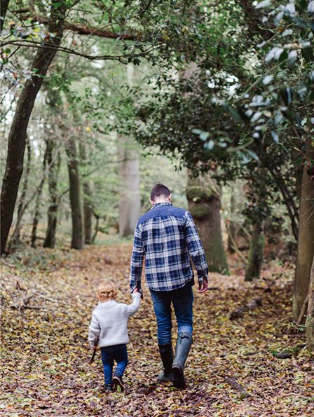 A parent and child walk hand in hand through a wooded area covered in fallen autumn leaves.