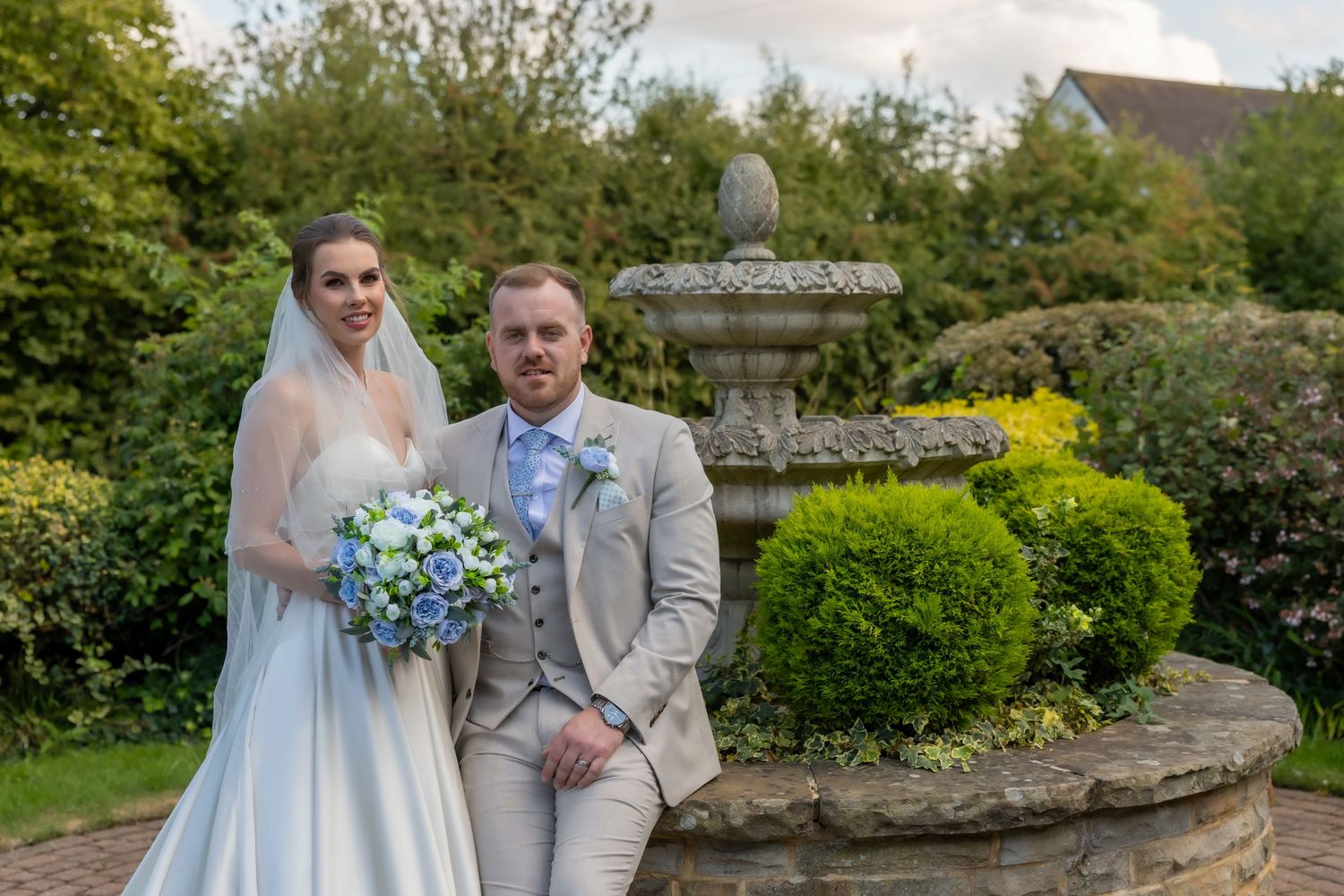 Wedding couple poses in a garden setting with blue floral bouquet and gray formal attire at an outdoor venue.