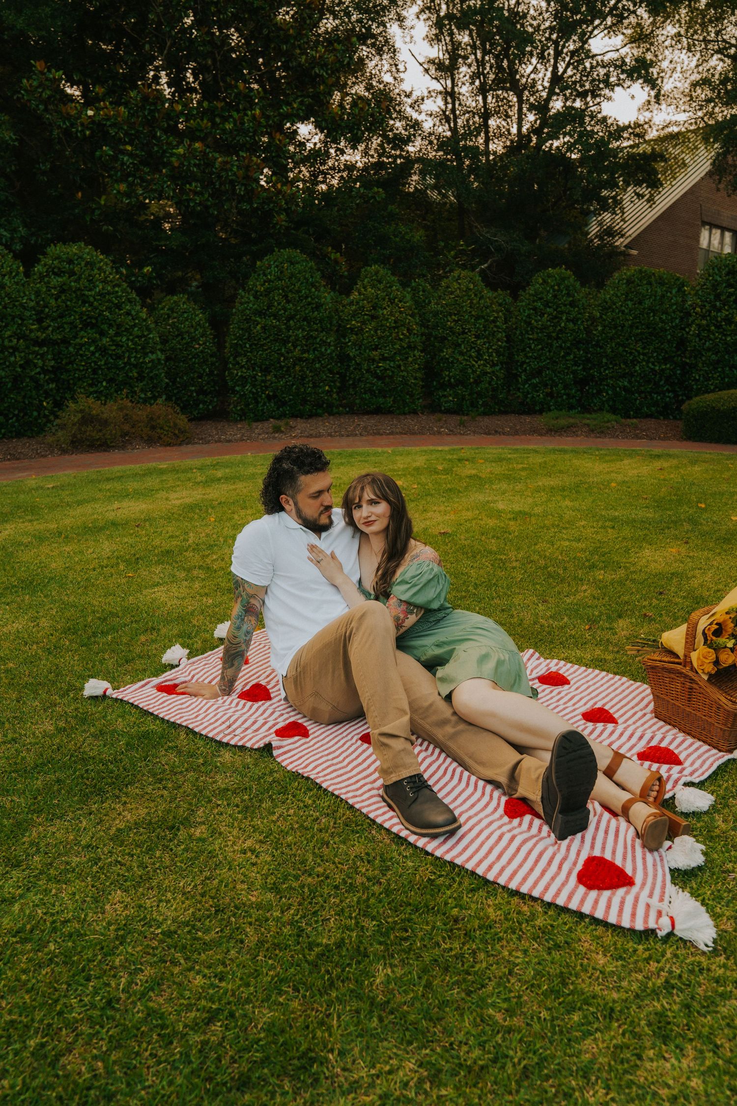 A couple relaxes on a red and white striped blanket during a romantic sunset picnic on a manicured lawn.