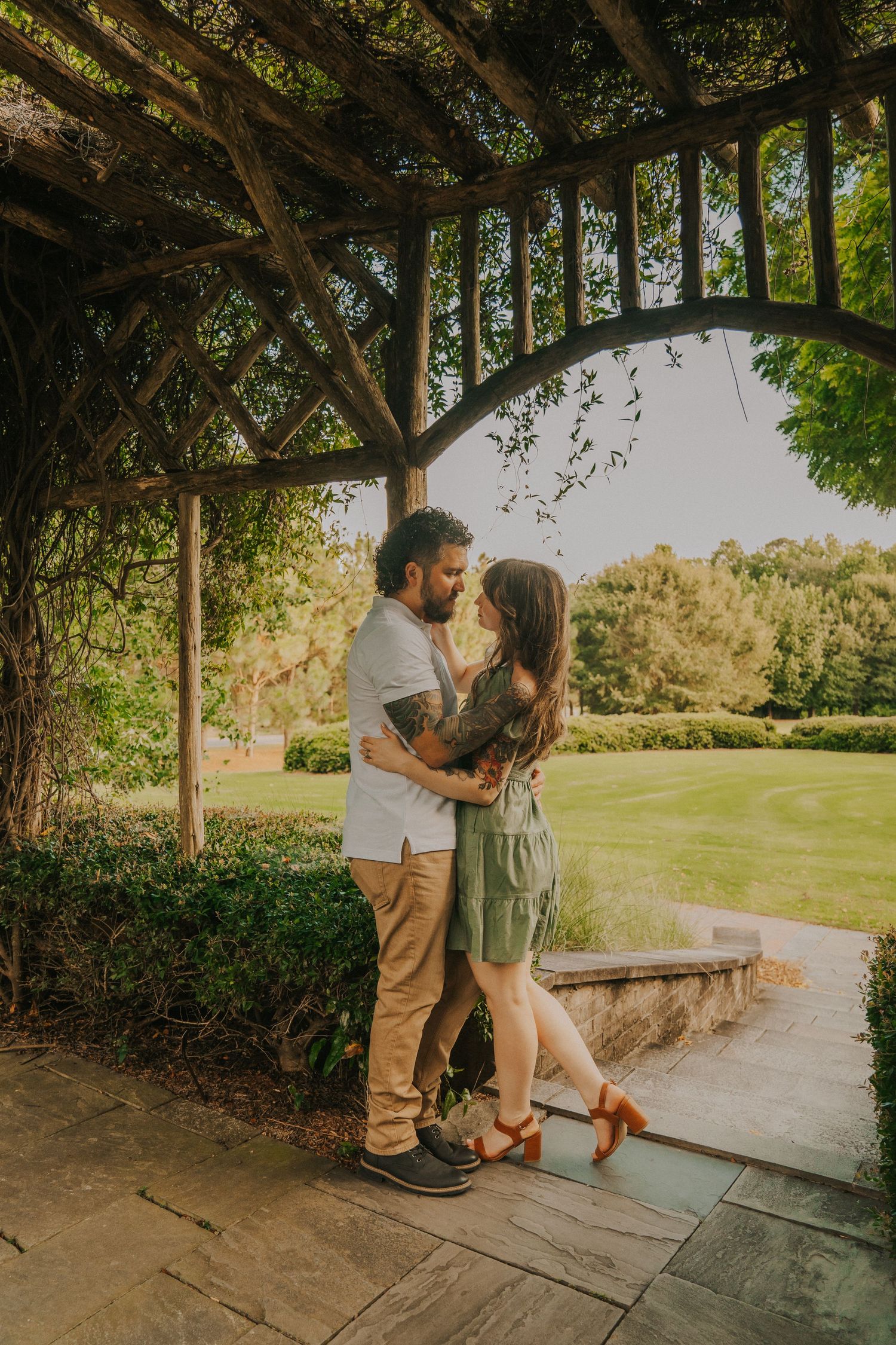 A romantic couple embraces under a vine-covered garden pergola during golden hour in a lush park setting.