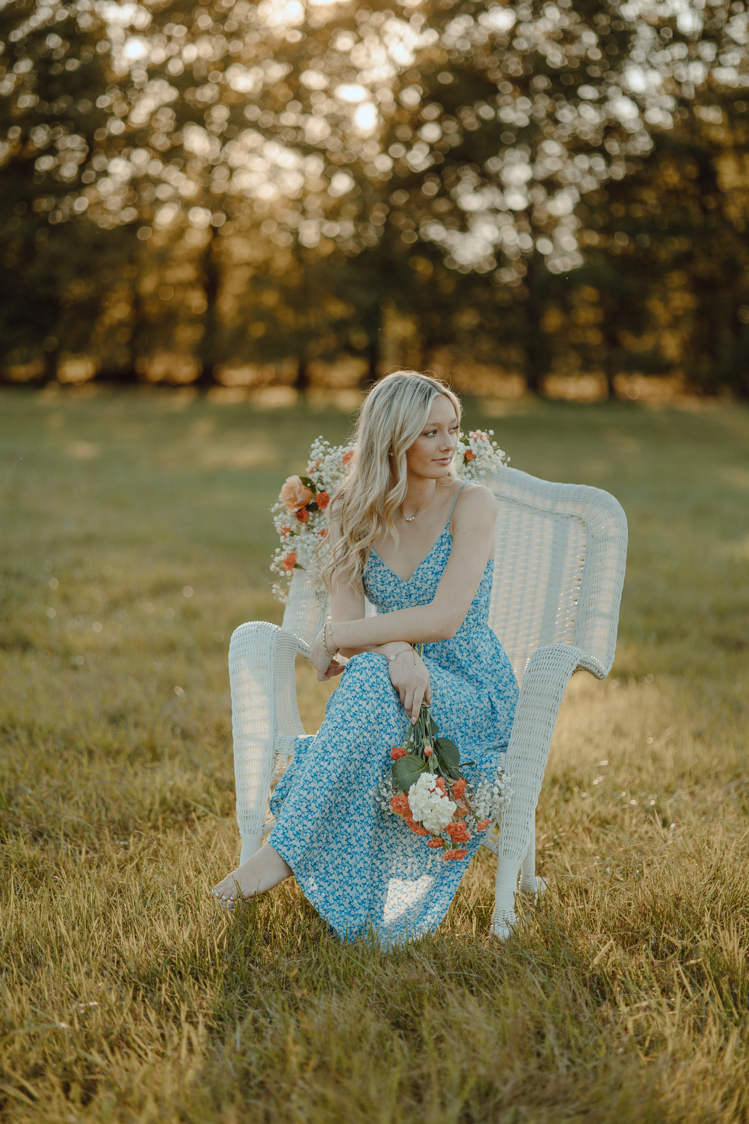 A person in a blue floral dress sits gracefully on a white vintage chair in a sunlit meadow at golden hour.