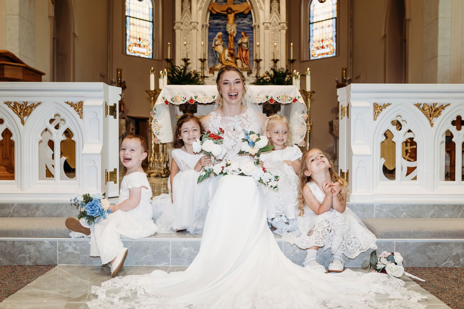A bride in a flowing white gown sits gracefully on a Catholic church floor surrounded by her flower girls posing for family portraits.