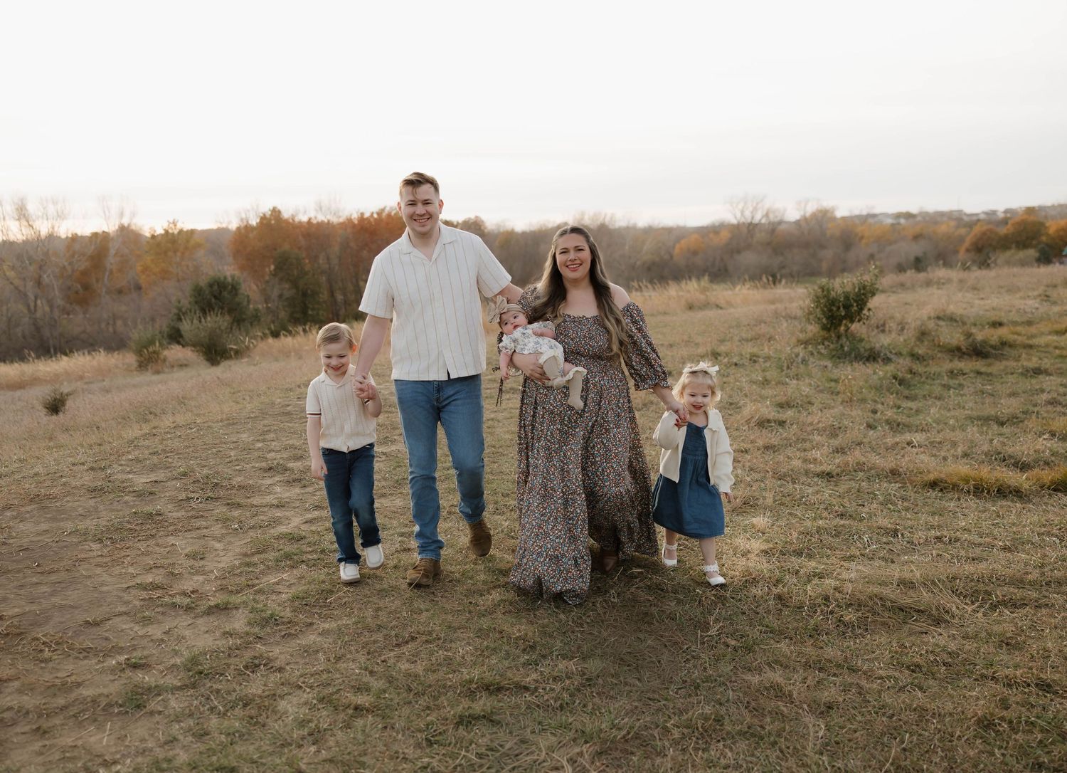 A family enjoying a tender moment together on a dirt path in a golden field with hills in the background at sunset.