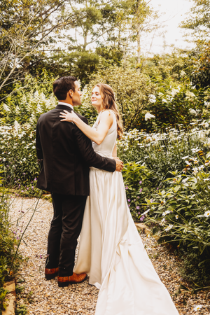 Couple in wedding attire walking along a gravel path through a garden setting.