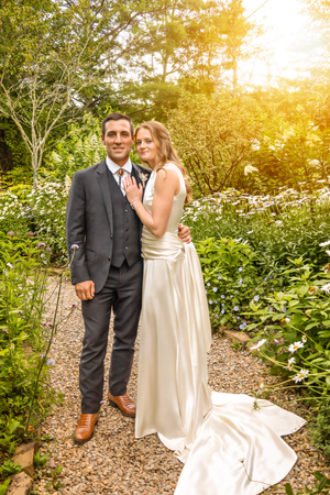 Wedding couple walking and posing on garden path with warm sunlight filtering through trees.