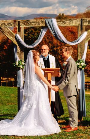 Wedding ceremony under white draped arch with fall foliage in background.