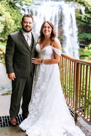 Newlyweds pose on wooden deck with stunning waterfall backdrop during their wedding photoshoot.