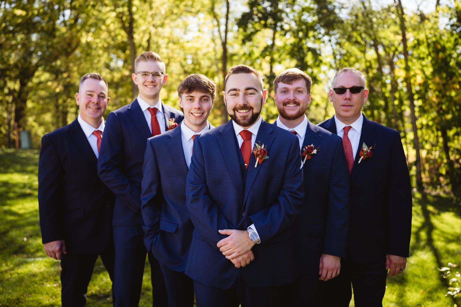 Groomsmen in navy blue suits and red ties pose together outdoors in a sunlit garden setting.