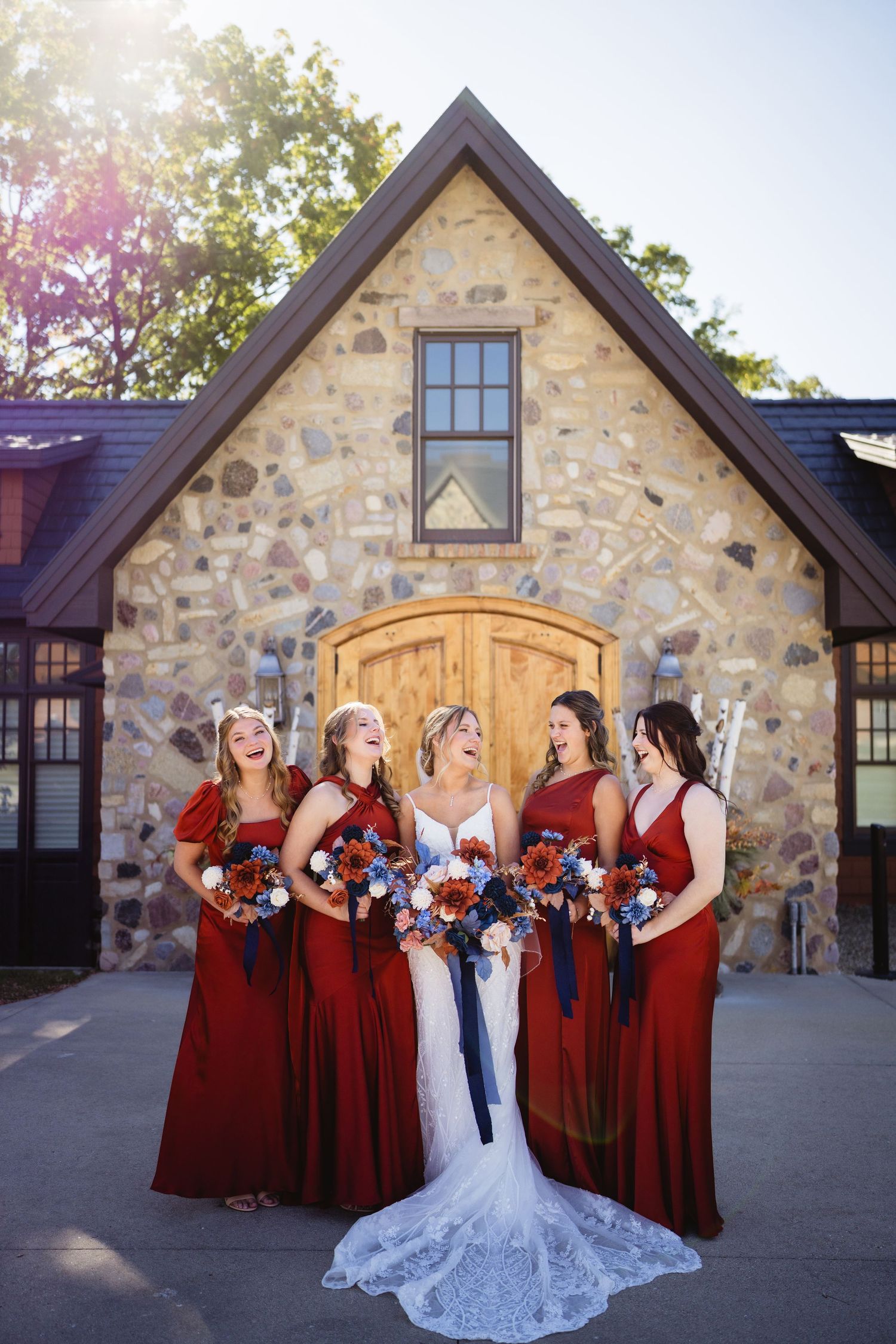 Wedding party in burgundy dresses and white gown pose together outside stone venue during golden hour celebration.