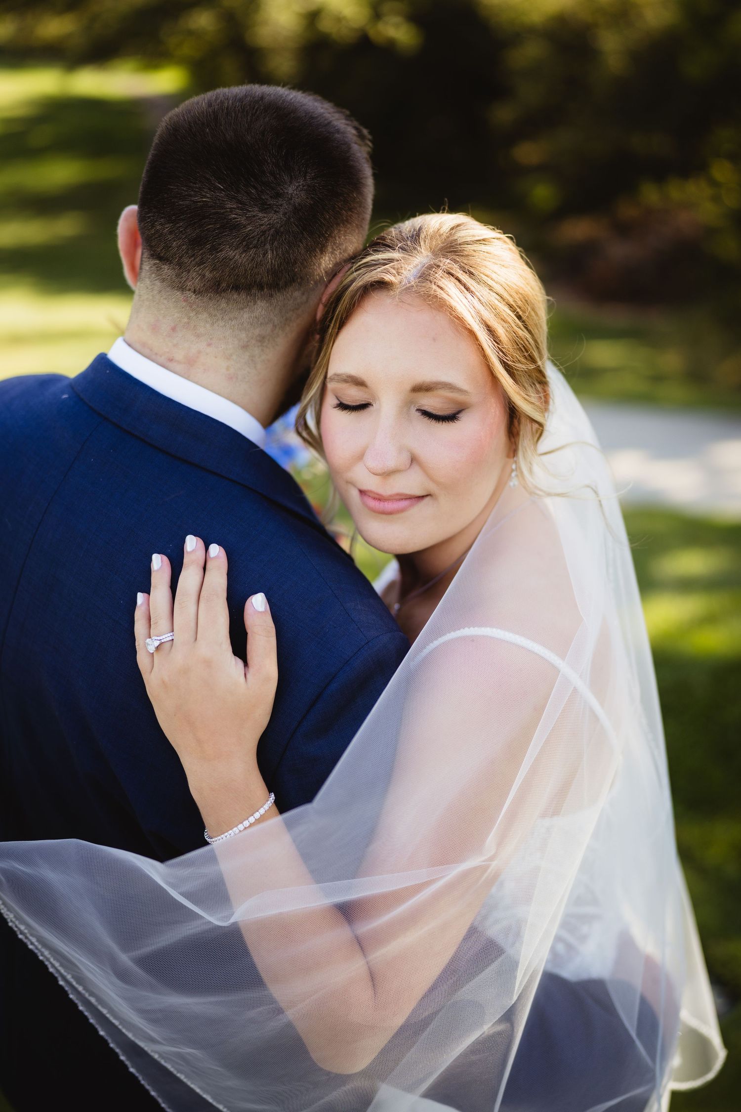 A tender moment between newlyweds as they embrace outdoors, with the bride wearing a flowing white veil and groom in navy suit.